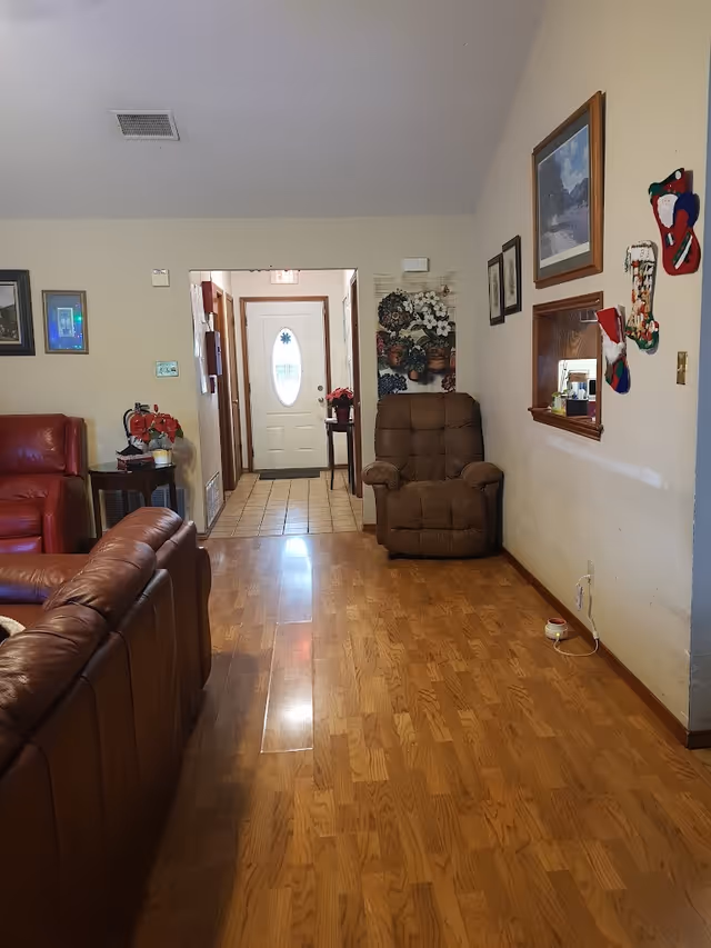 Living room with hardwood floors, red leather sofas, a brown recliner, wall artwork and a view toward an entry door.
