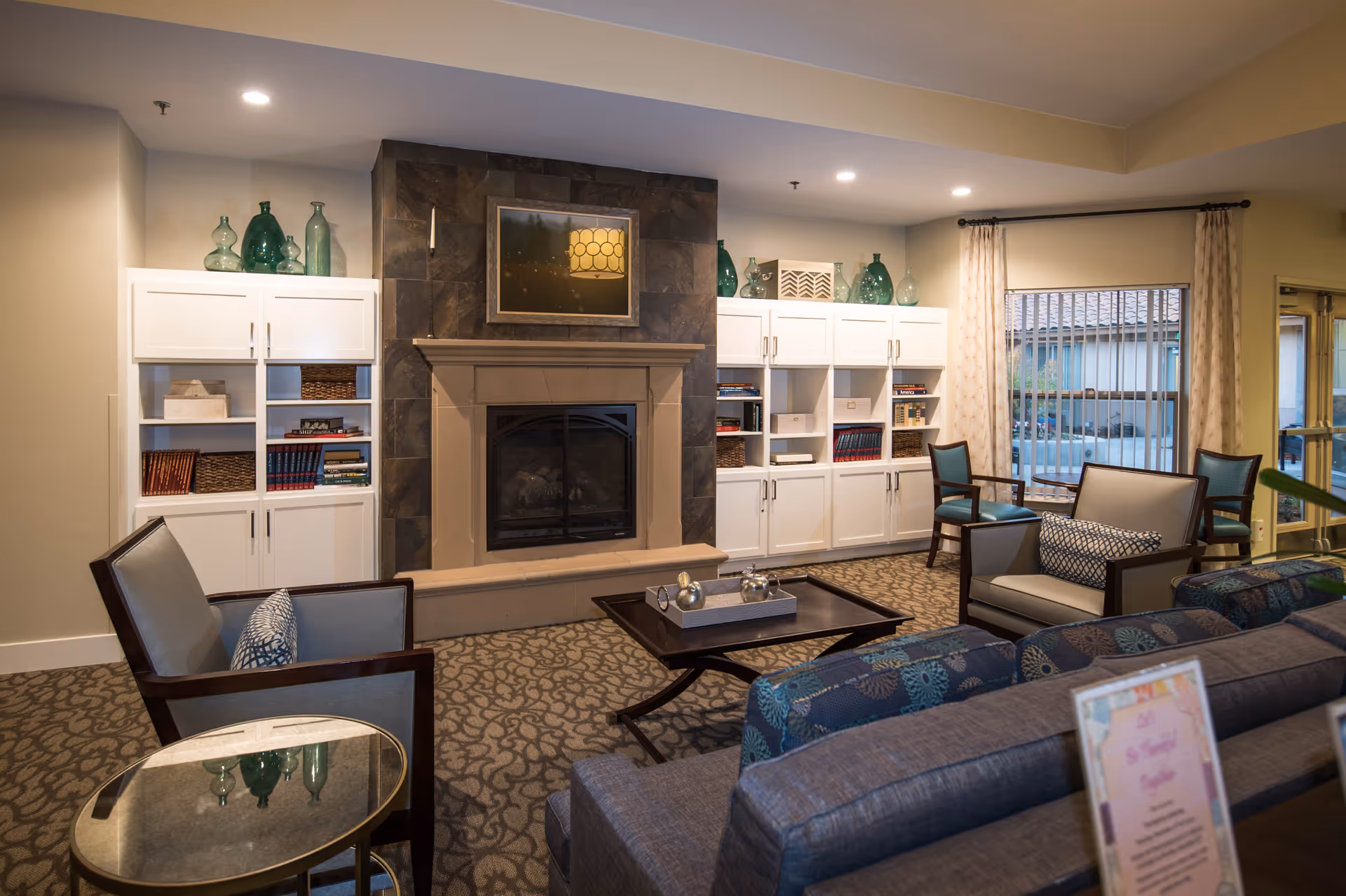 A cozy living room area in Meadow Oaks of Roseville featuring a stone fireplace with a framed picture above it, white built-in cabinets with decorative glass bottles and books, several upholstered chairs and sofas arranged around a coffee table, patterned carpet, and large windows with curtains letting in natural light.