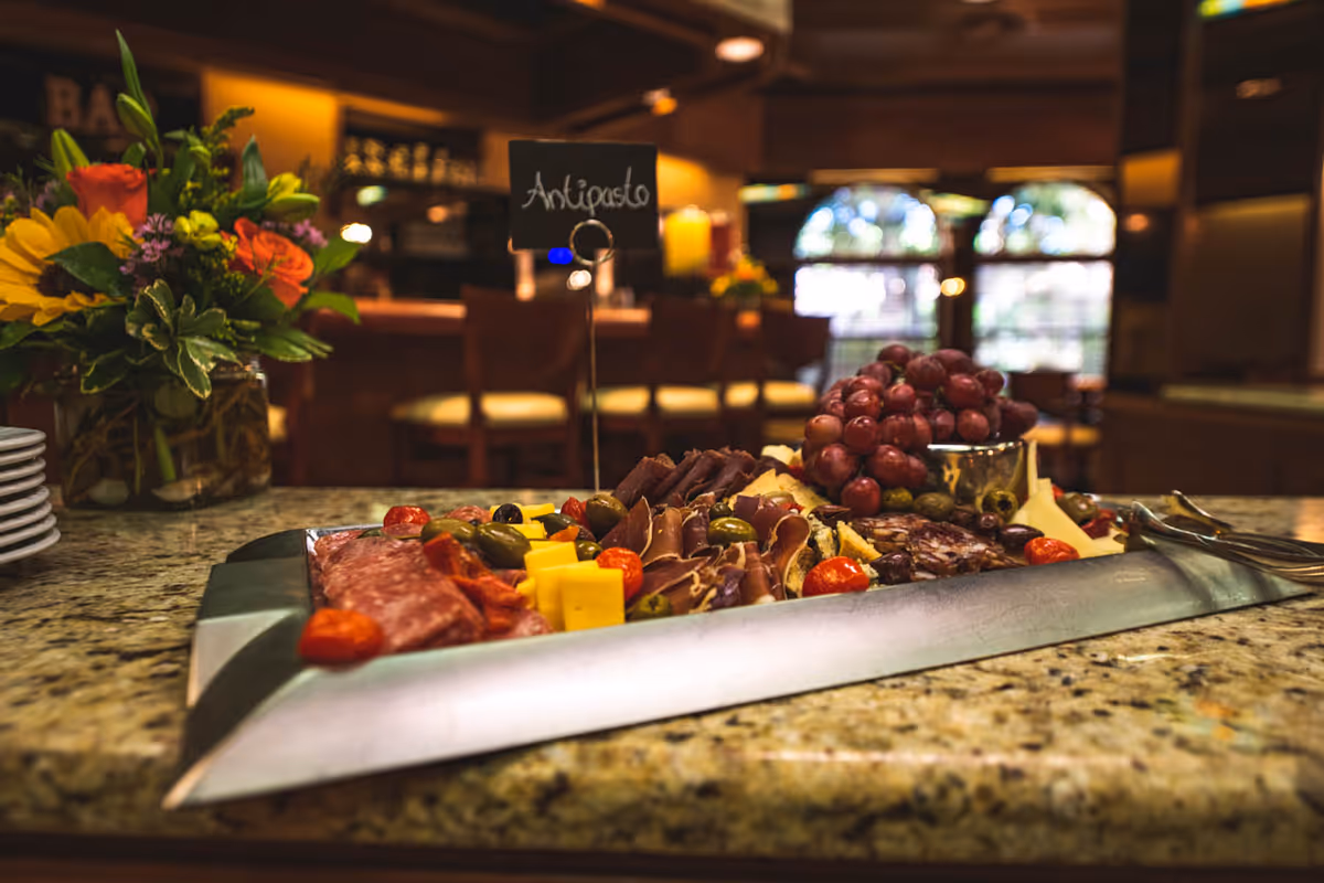 A close-up view of an antipasto platter with assorted meats, cheeses, olives, cherry tomatoes, and grapes on a granite countertop in a warmly lit dining area with wooden chairs and floral arrangements in the background.