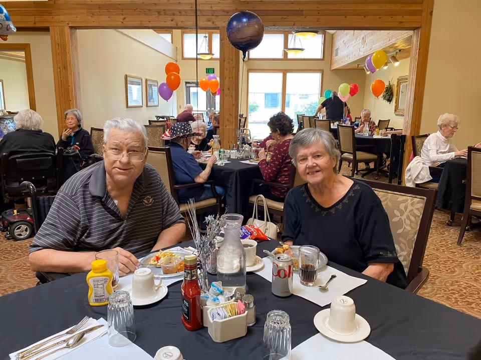 Two elderly people sitting at a dining table in a retirement community dining room, enjoying a meal with drinks and condiments on the table. Other elderly residents are seated at tables in the background, with colorful balloons decorating the room.