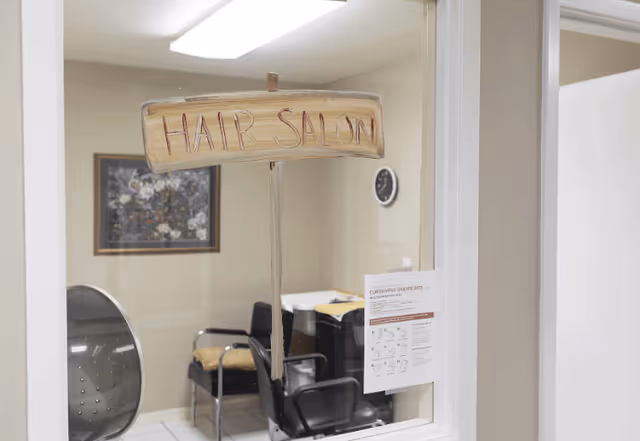 View through a window into a hair salon room with a wooden sign reading 'HAIR SALON' hanging in the window. Inside the room are black salon chairs, a framed floral picture on the wall, a clock, and a posted notice about coronavirus disease 2019.