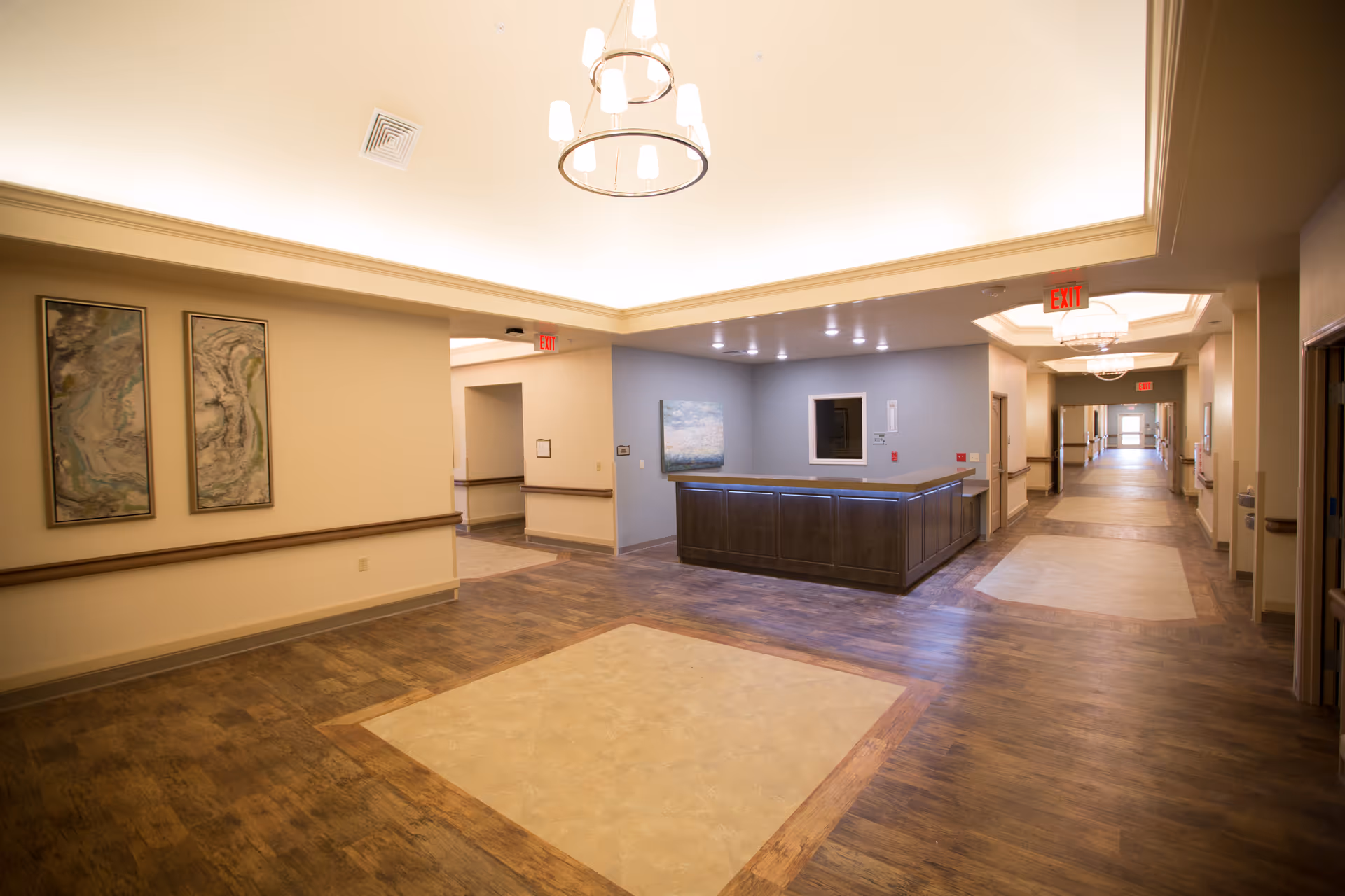 A spacious, well-lit hallway in a healthcare facility with wood-patterned flooring and beige walls. There is a reception desk with a blue-gray wall behind it, decorated with a painting. The ceiling features recessed lighting and modern chandeliers. Exit signs are visible above doorways along the hallway.