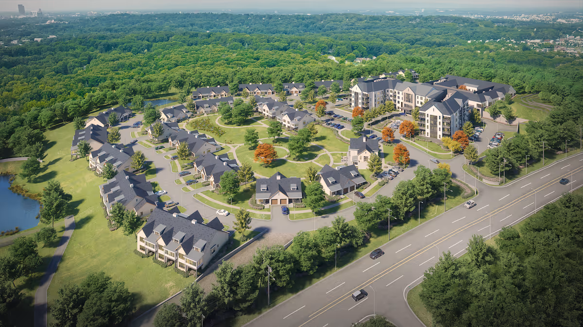 Aerial view of The Providence Senior Living facility showing multiple residential buildings arranged in a circular pattern around green lawns and trees, with a main road in the foreground and dense forest in the background.