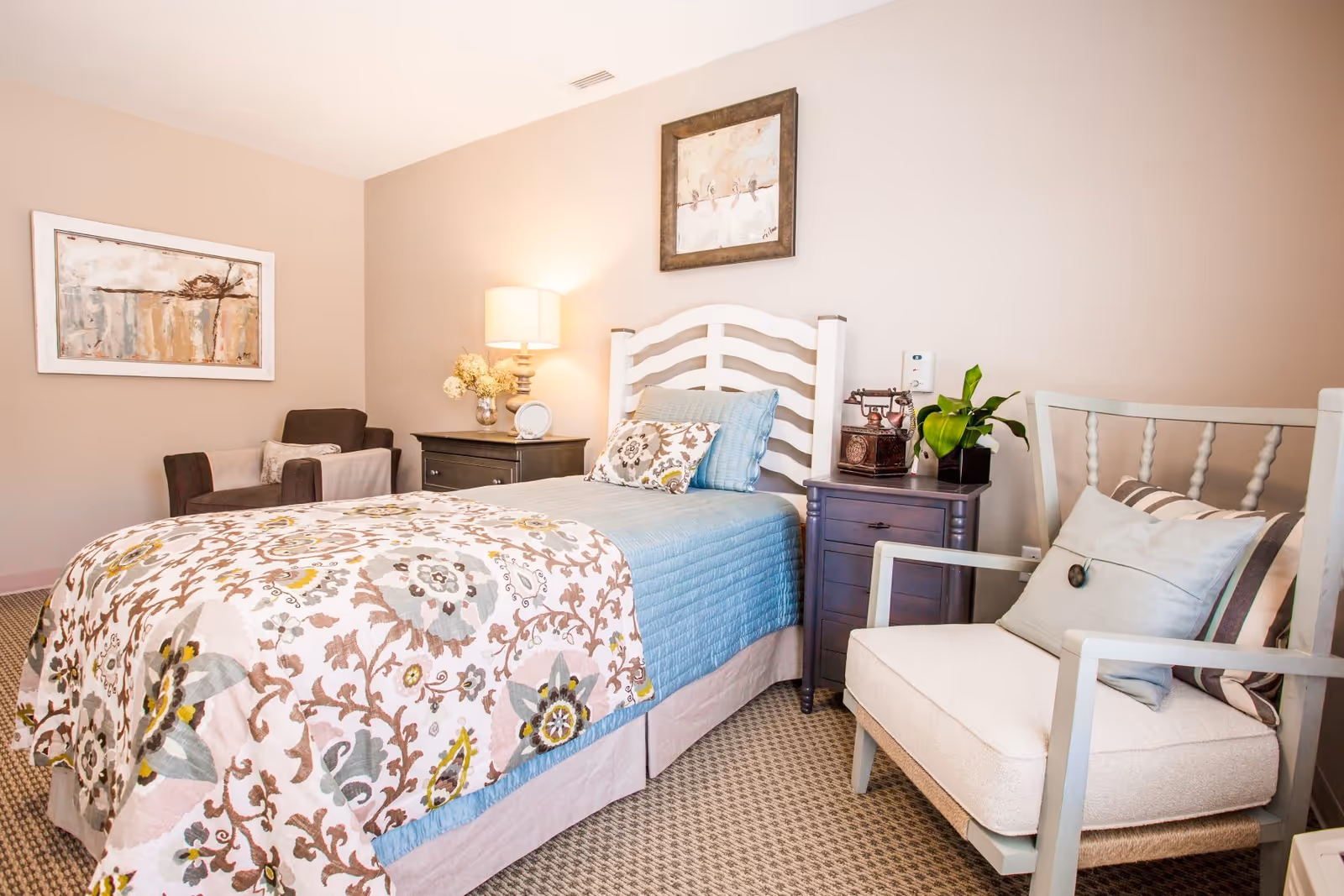 A cozy furnished bedroom with a single bed, nightstands, armchairs, a lamp, and wall art.