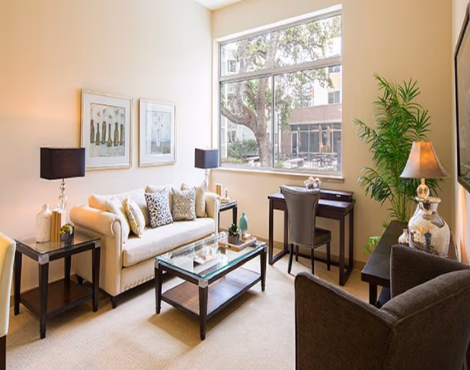 Bright senior living lounge with sofa, chairs, glass coffee table, and large window.