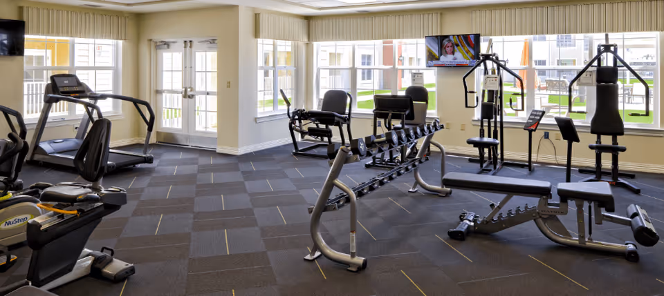 Well-lit fitness room with treadmills, weight benches, strength machines, and dumbbells in front of large windows.