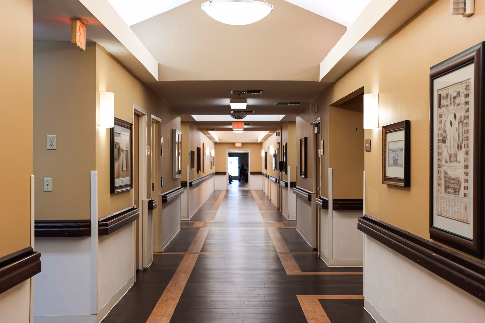A long, well-lit hallway in a senior living facility with beige walls, dark wood handrails, framed artwork on the walls, and multiple doors leading to rooms on either side. The floor has a patterned design with dark and light wood tones, and exit signs are visible at both ends of the corridor.