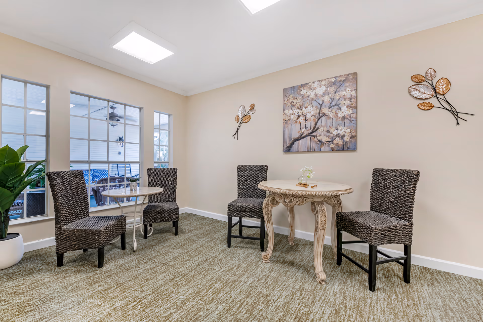 A small seating area with two round tables and four woven chairs in a room with beige walls and carpeted floor. The wall is decorated with a painting of a flowering tree and two metal leaf wall hangings. Large windows reveal an adjacent covered outdoor area with additional seating and a ceiling fan.