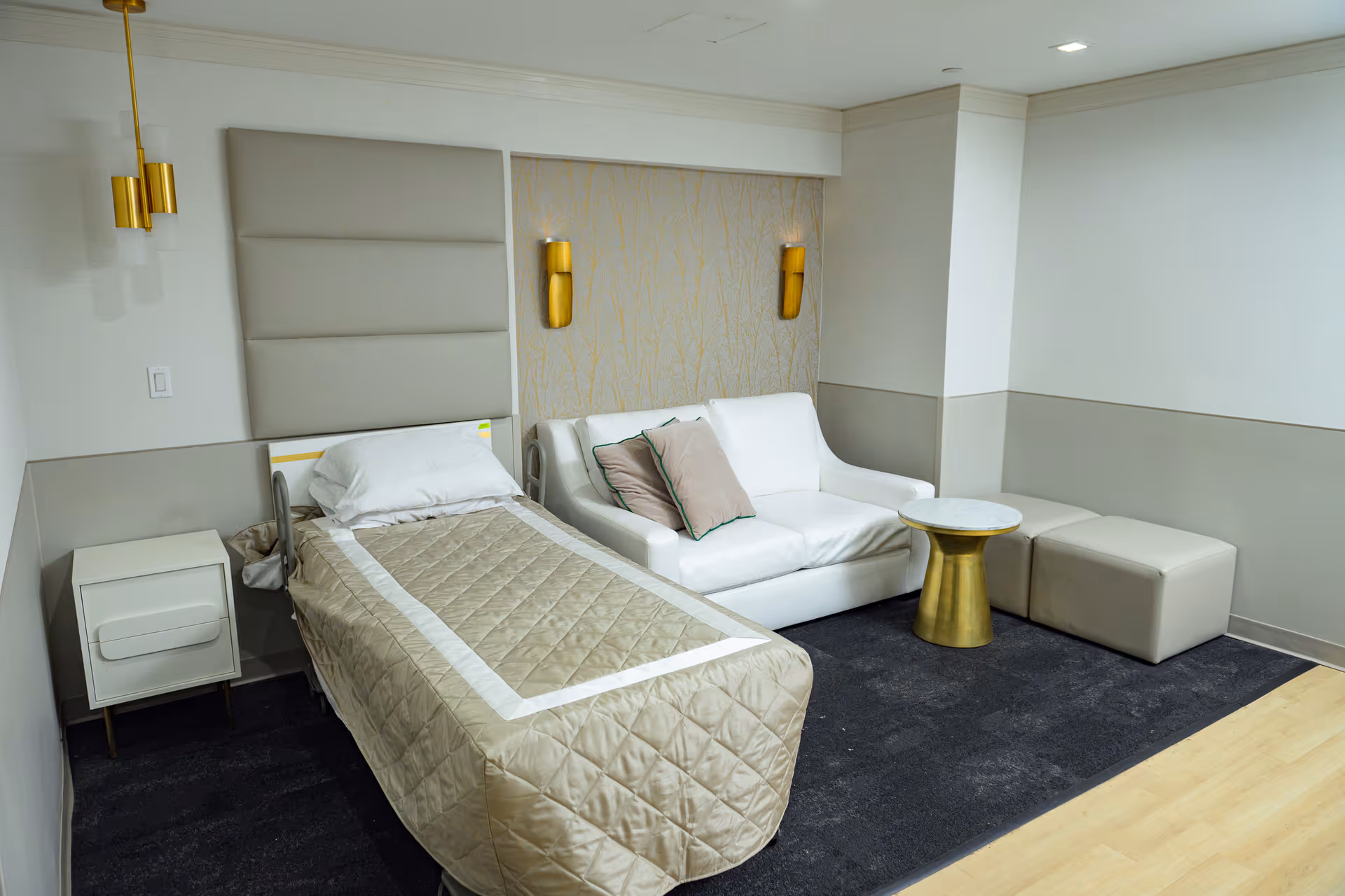 A neatly arranged room with a single bed covered with a beige quilt, a white pillow, and a padded headboard. Next to the bed is a small white nightstand. Adjacent to the bed is a white loveseat with two beige pillows, a small round gold and white table, and two beige ottomans. The room has light-colored walls with gold wall sconces and a dark carpeted floor with a light wood border.