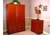 A wooden armoire and matching three-drawer dresser with a lamp and wall decorations against a pink-painted wall.