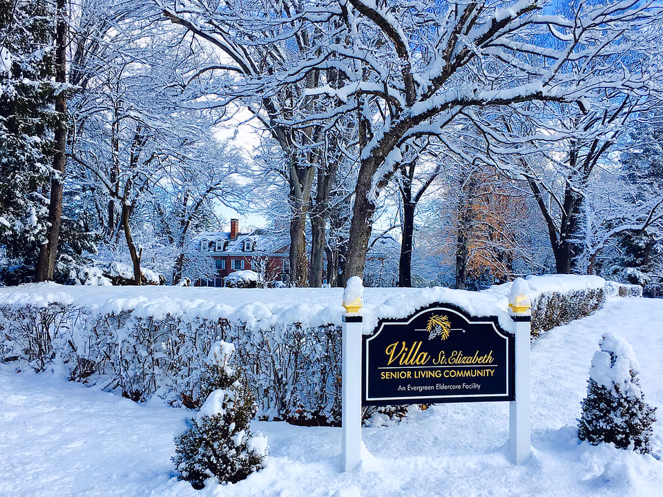 Snow-covered grounds and trees with a 'Villa St. Elizabeth Senior Living Community' sign in front of a brick residence.