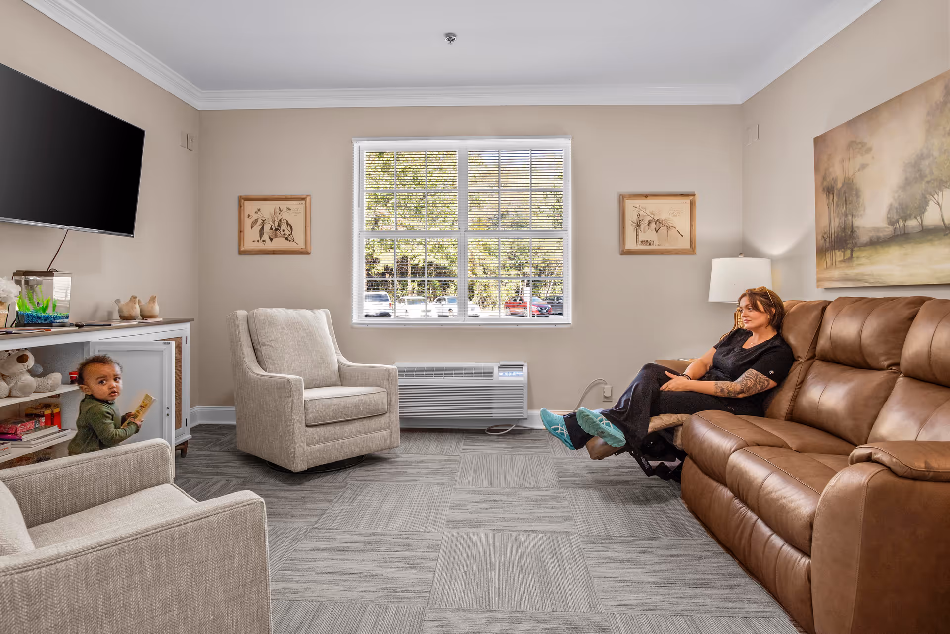 A cozy living room with beige walls and gray carpet tiles. There is a large window with white blinds letting in natural light. A woman is sitting on a brown leather reclining sofa on the right side, and a young child is standing near a white cabinet on the left. The room also features two beige armchairs, a wall-mounted flat-screen TV, framed artwork on the walls, and a table lamp beside the sofa.