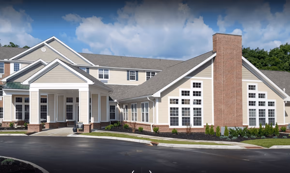 Exterior view of The Ashford of Mt. Washington senior living facility showing a large building with beige siding, multiple windows, a prominent brick chimney, and a covered entrance. The sky is partly cloudy and there is a paved driveway in front.