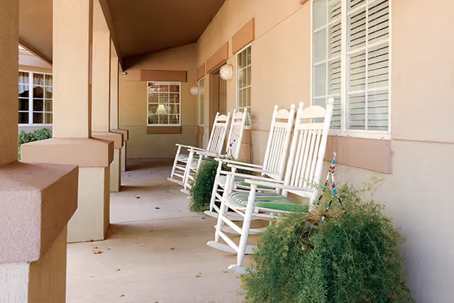 Covered front porch of an assisted-living building with white rocking chairs and potted greenery along a beige exterior.