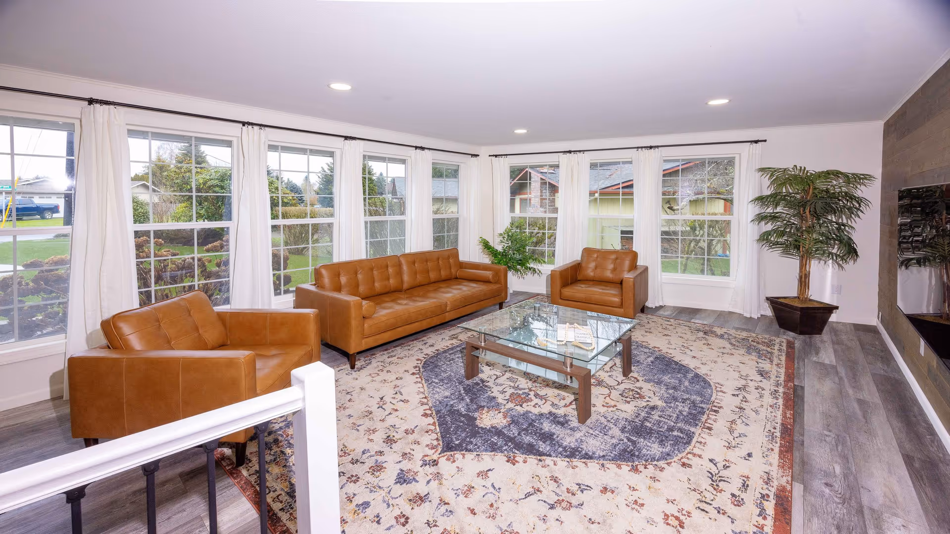 A bright living room with large windows covered by white curtains, featuring a brown leather sofa, two matching armchairs, a glass coffee table on a patterned area rug, and a potted plant in the corner.