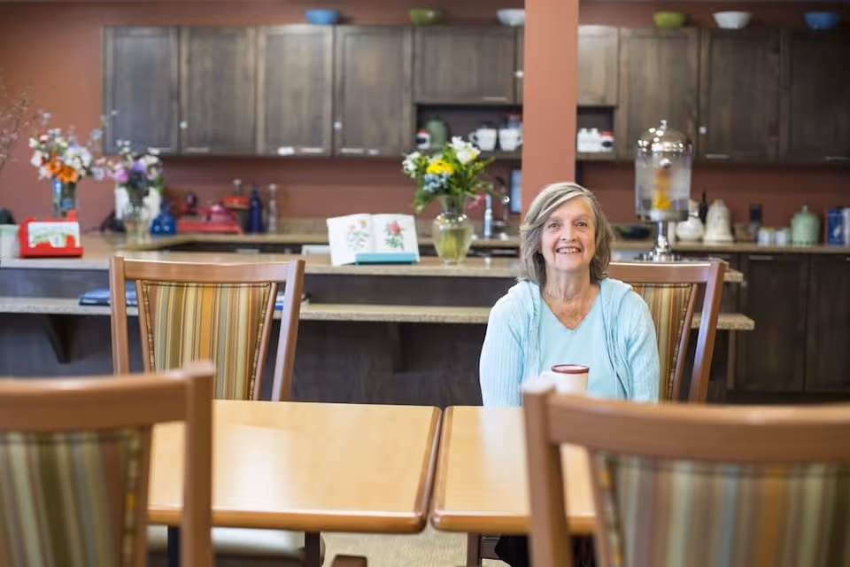 An elderly woman with gray hair wearing a light blue sweater sits at a wooden table with striped chairs in a kitchen or dining area. Behind her, there are dark wooden cabinets, a countertop with flowers in vases, a water dispenser, and various kitchen items.