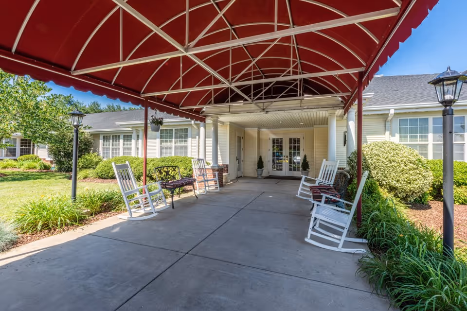 Covered entrance area of a senior living facility with a red canopy overhead, white rocking chairs and a bench along the sides, and a concrete walkway leading to double glass doors. The building exterior is light-colored with large windows and surrounded by green bushes and plants under a clear blue sky.
