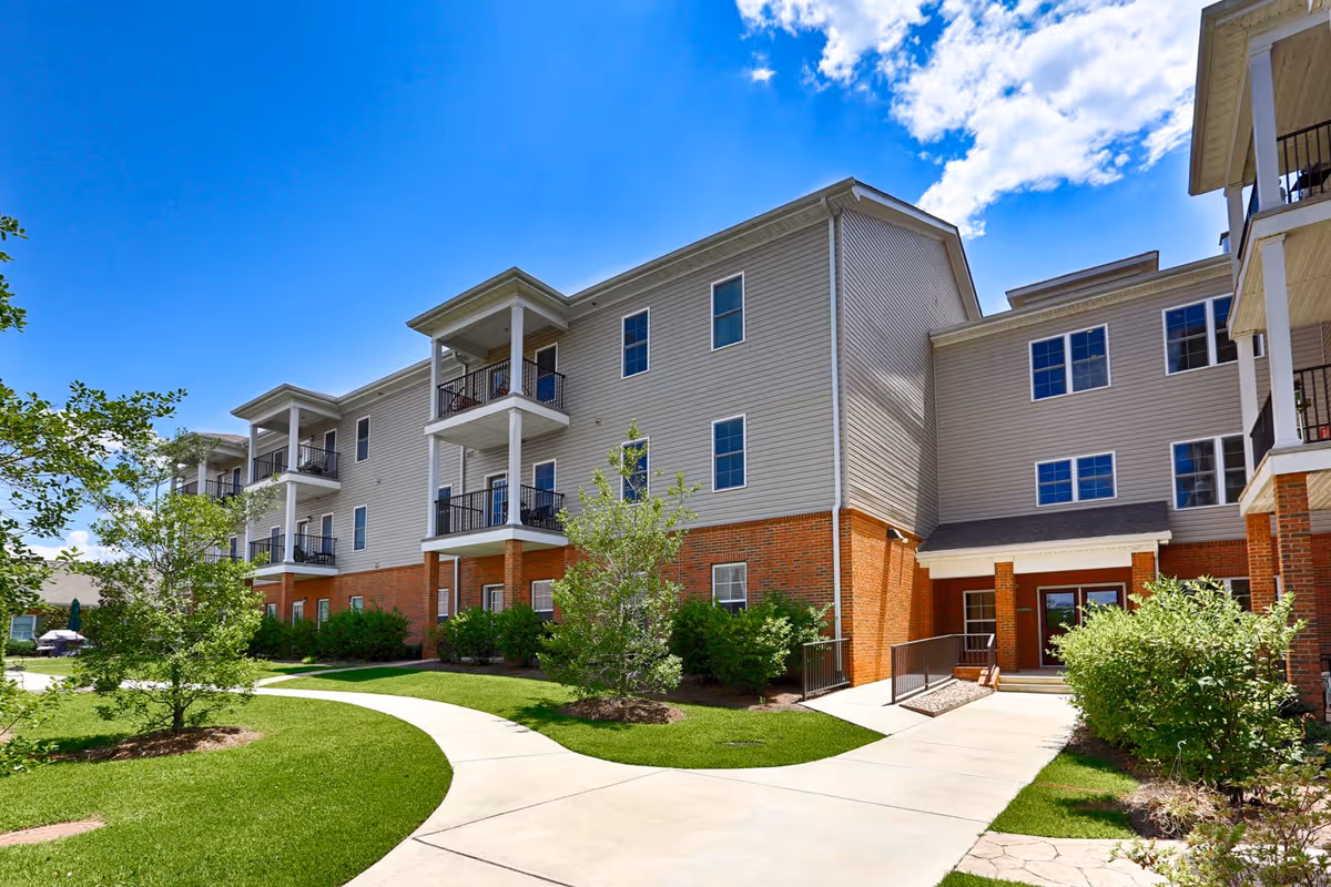 Exterior view of a three-story residential building with beige siding and red brick lower walls, featuring balconies and surrounded by green lawns, shrubs, and trees under a blue sky with some clouds.