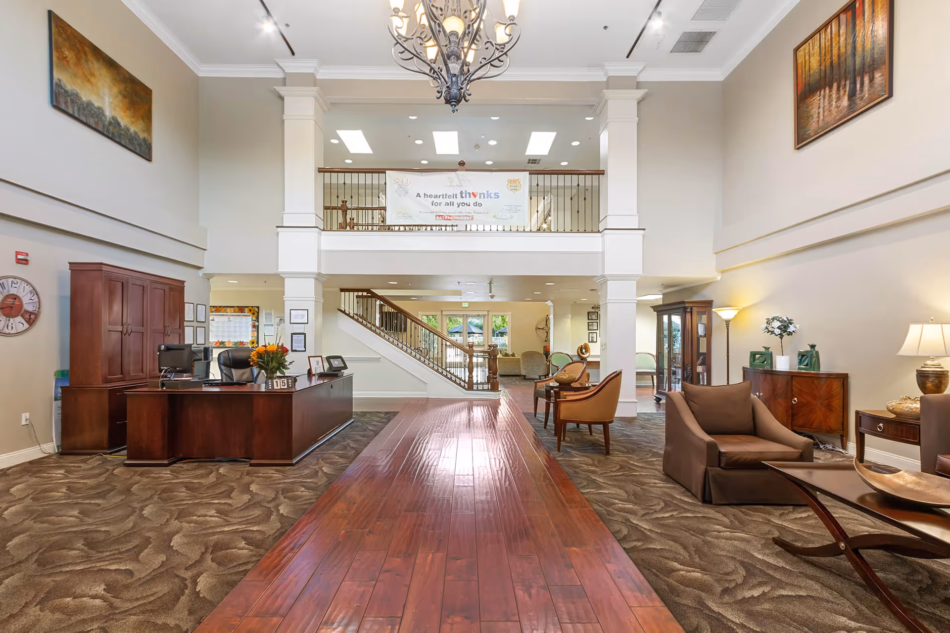 Spacious two-story lobby with a reception desk, seating area, central wooden walkway, staircase and chandelier.