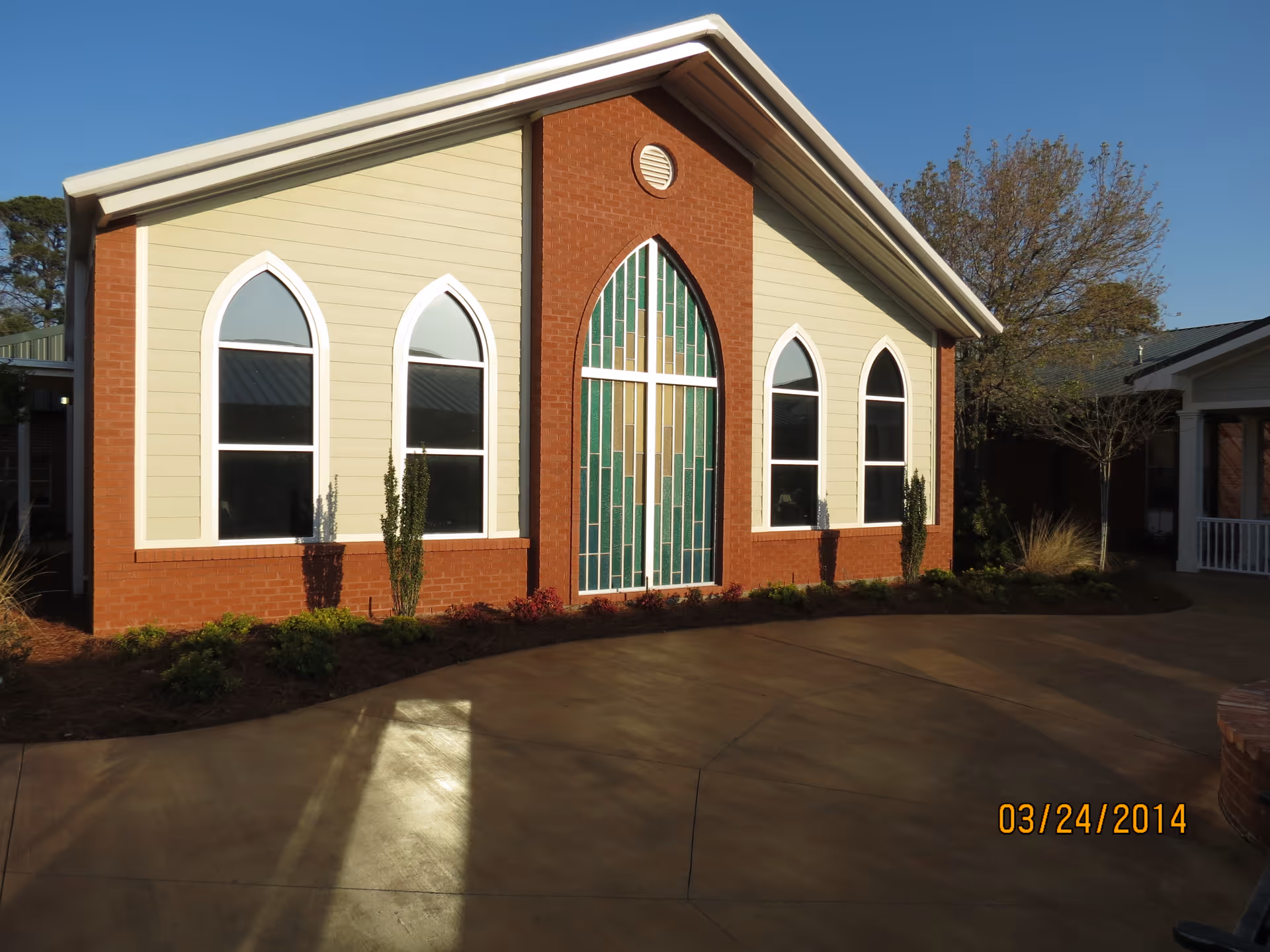 Exterior view of a building with a brick and light-colored siding facade featuring five tall, narrow arched windows. The central window is a stained glass design with a cross. There are small shrubs and plants along the base of the building, and a paved area in front. The sky is clear and blue.