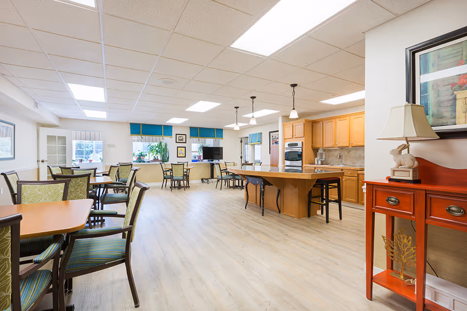 A bright and spacious senior living dining area with several wooden tables and green cushioned chairs. The room features a kitchen area with wooden cabinets, a large island with stools, and pendant lighting. There are windows with blue valances, a TV on a stand, and a red side table with a lamp and decorative items.