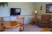 A cozy interior room with a wooden dining table and chairs, a brown upholstered armchair, a side table with a lamp, a wall-mounted flat screen TV, and framed artwork on the wall.
