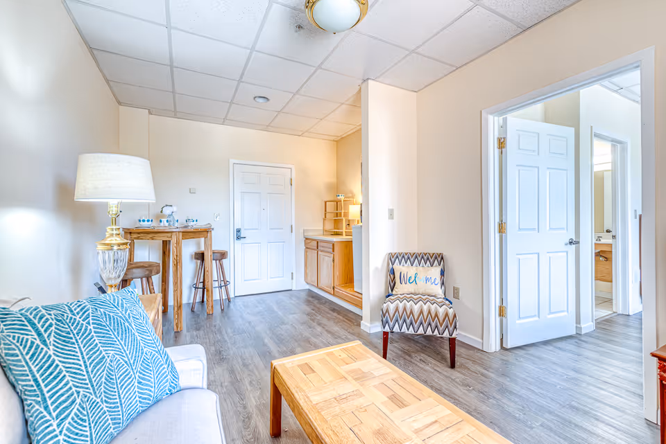 Bright and clean living room area in an active adult living facility featuring a sofa with blue patterned pillows, a wooden coffee table, a lamp on a side table, a small wooden dining table with two stools, and a patterned chair with a 'Welcome' pillow. The room has light-colored walls, wood flooring, and a white door leading to another room with a visible bathroom.