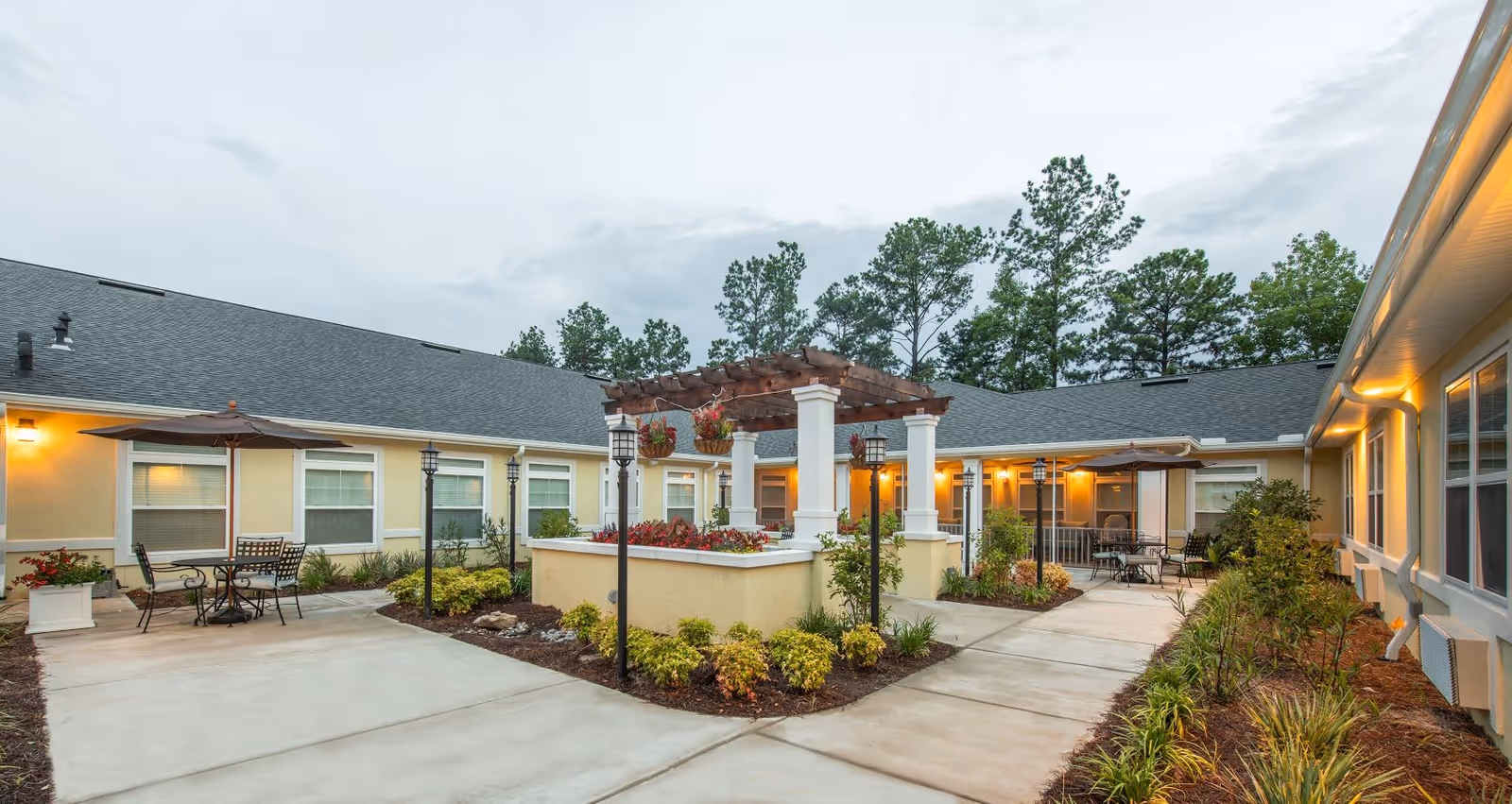 Outdoor courtyard area at The Addison of West Ashley featuring a central pergola with hanging flower baskets, surrounded by paved walkways, garden beds with shrubs, and multiple seating areas with tables and umbrellas. The building exterior is light yellow with white trim and lit wall sconces, with tall trees in the background under a cloudy sky.