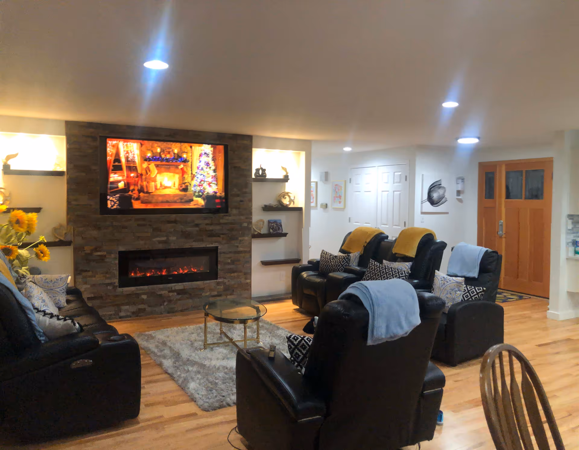 A cozy living room with several black leather recliners, each with patterned pillows and colorful throws. A stone fireplace with an electric fire insert is centered on one wall, above which is a mounted flat-screen TV displaying a festive scene with a Christmas tree and fireplace. The room has wooden flooring, recessed ceiling lights, built-in shelves with decorative items, and a wooden front door in the background.