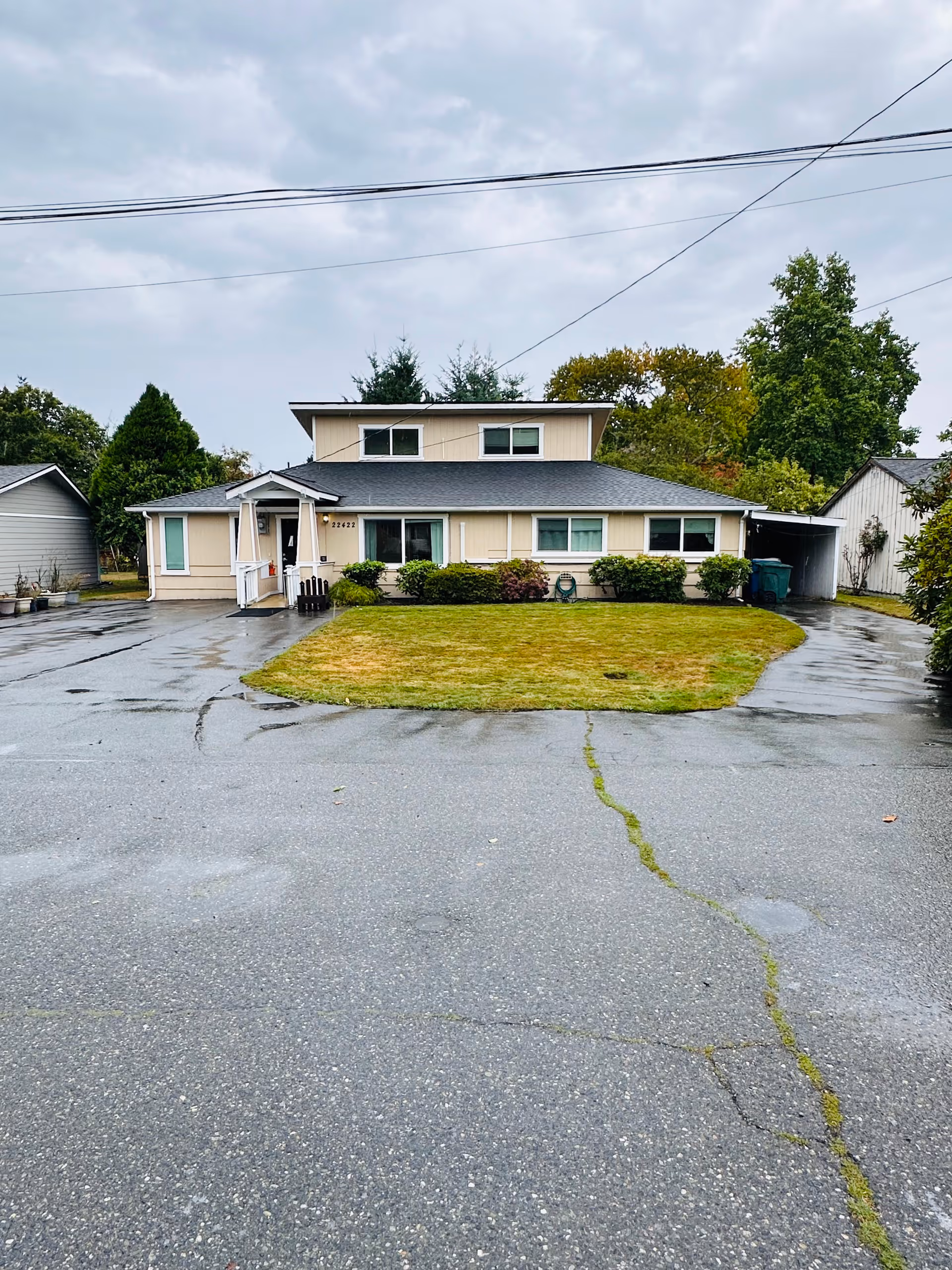 Front exterior view of a single-story residential building with beige siding and a dark roof, surrounded by a wet driveway and a small lawn with shrubs. The sky is overcast and there are trees in the background.