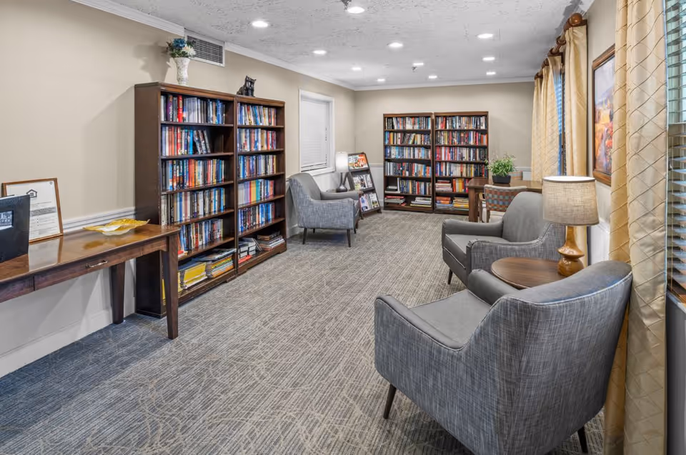 A cozy reading room with carpeted floor, beige walls, and ceiling lights. The room features two tall wooden bookshelves filled with books, three gray upholstered armchairs, a small round wooden table with a lamp, a wooden desk with papers and a framed certificate, and a window with beige curtains. There is also a small plant on a table near the back bookshelf.