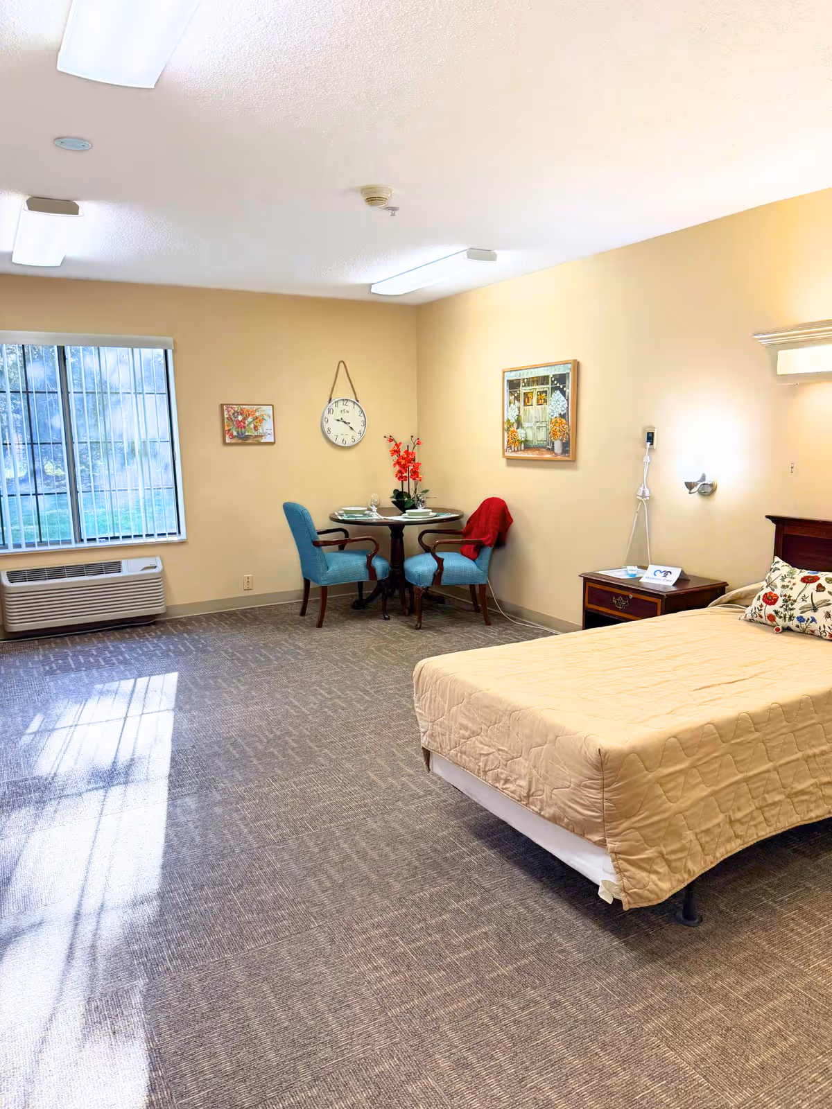 A bright and clean assisted living bedroom with a single bed covered in a beige quilt, a wooden nightstand with a lamp and phone, two blue upholstered chairs around a small round table with a flower vase, wall clock, and framed artwork. A large window with vertical blinds lets in natural light, and the room has beige walls and carpeted floor.