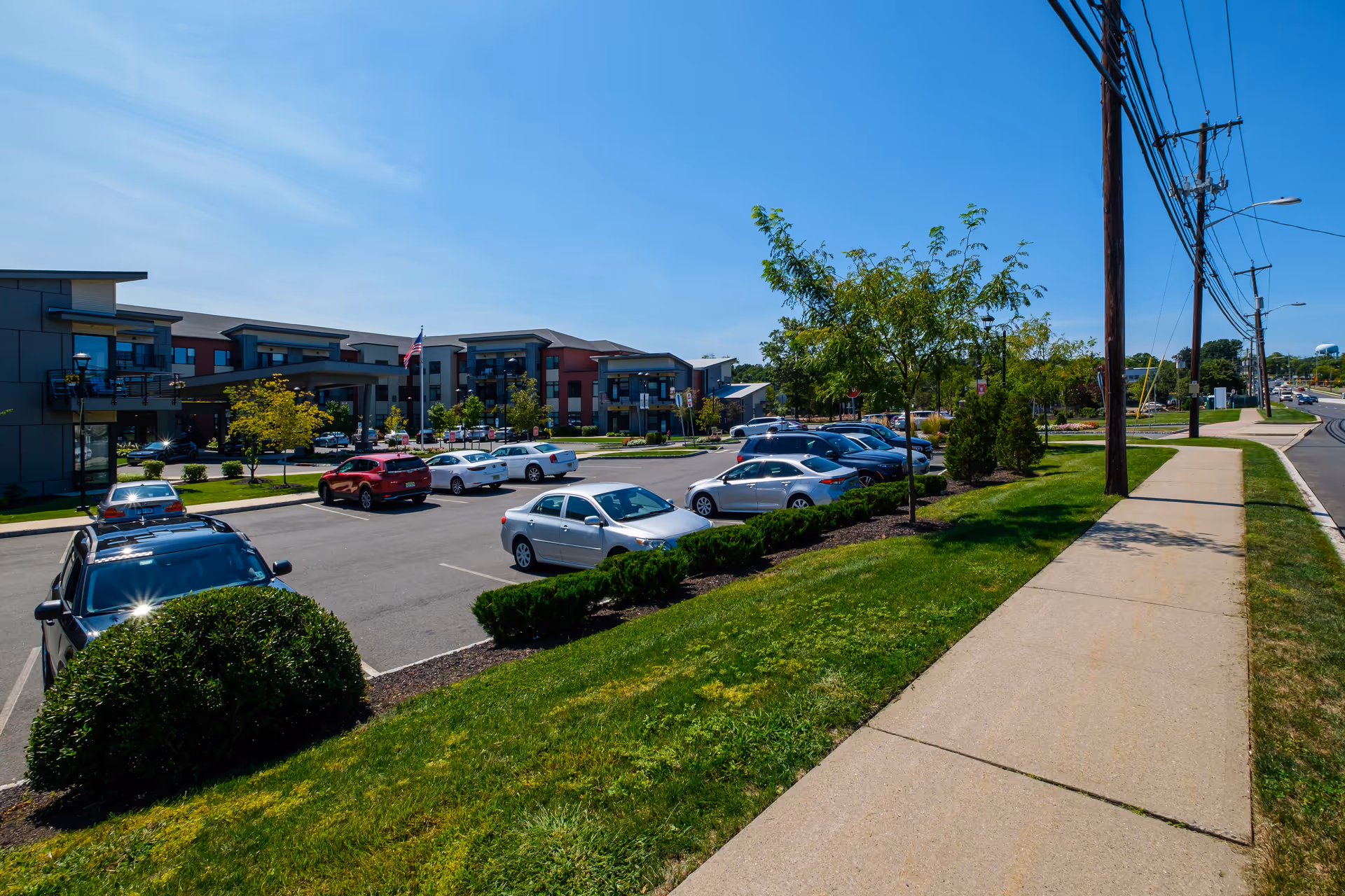View of a senior living facility parking lot with several parked cars, green grass, small trees, and a sidewalk alongside a road with utility poles under a clear blue sky.