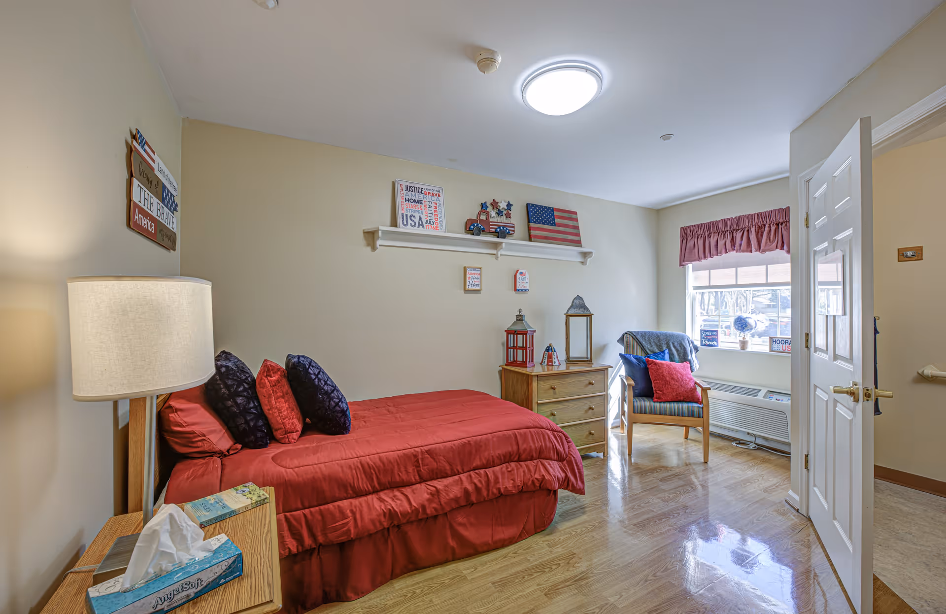 A cozy bedroom with a single bed covered in a red comforter and multiple pillows. There is a wooden nightstand with a lamp and a box of tissues next to the bed. On the wall above the bed, there are patriotic decorations including an American flag and themed signs. A wooden dresser with decorative lanterns sits against the wall, and a chair with cushions is placed near a window with a red valance. The room has wooden flooring and a white door partially open to an adjacent room.