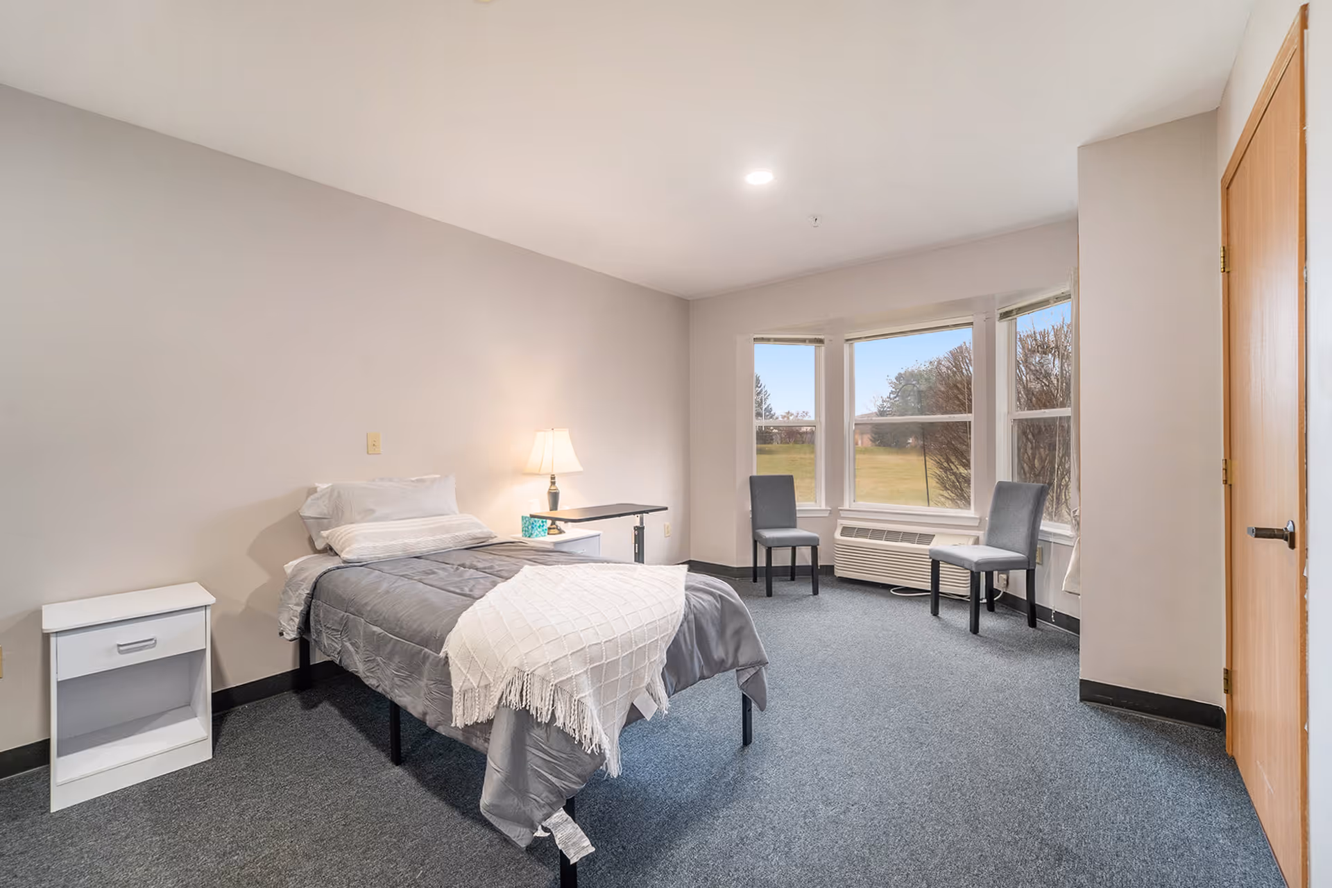 Simple furnished bedroom with a single bed, nightstand and lamp, two chairs by a bay window overlooking grassy grounds.