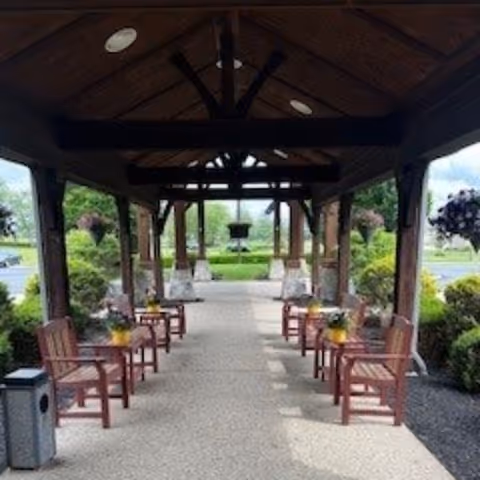 Covered outdoor walkway with wooden benches on both sides, potted plants on the benches, and greenery surrounding the area under a wooden roof structure.