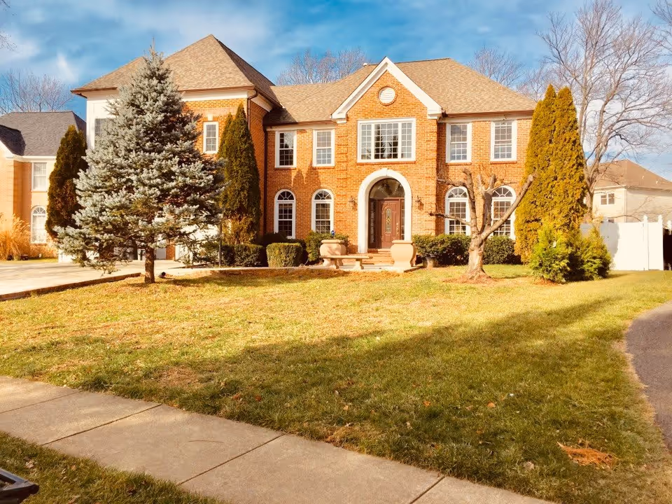 Front exterior view of a large two-story brick house with multiple windows, a central arched doorway, and a well-maintained lawn with trees and shrubs under a partly cloudy sky.