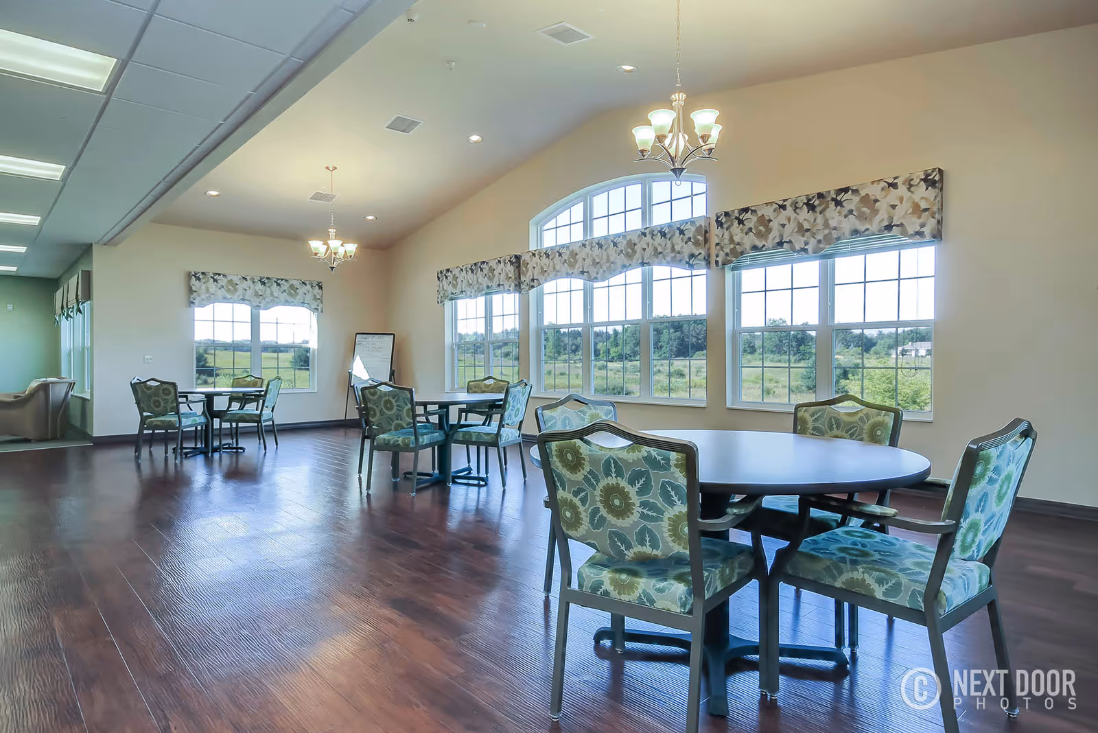 A spacious dining room with several round tables and floral-patterned chairs. Large windows with floral valances allow natural light to fill the room, offering a view of green fields outside. The room has wooden flooring and two chandeliers hanging from the ceiling.