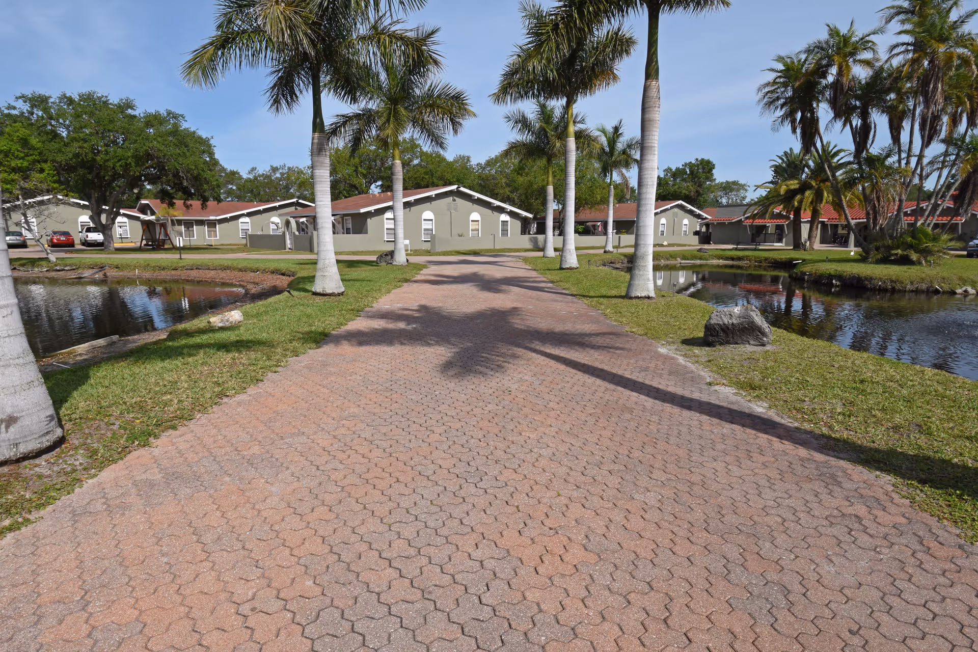 Paved walkway lined with palm trees leading to single-story buildings with red roofs, surrounded by small ponds and green grass under a clear blue sky.