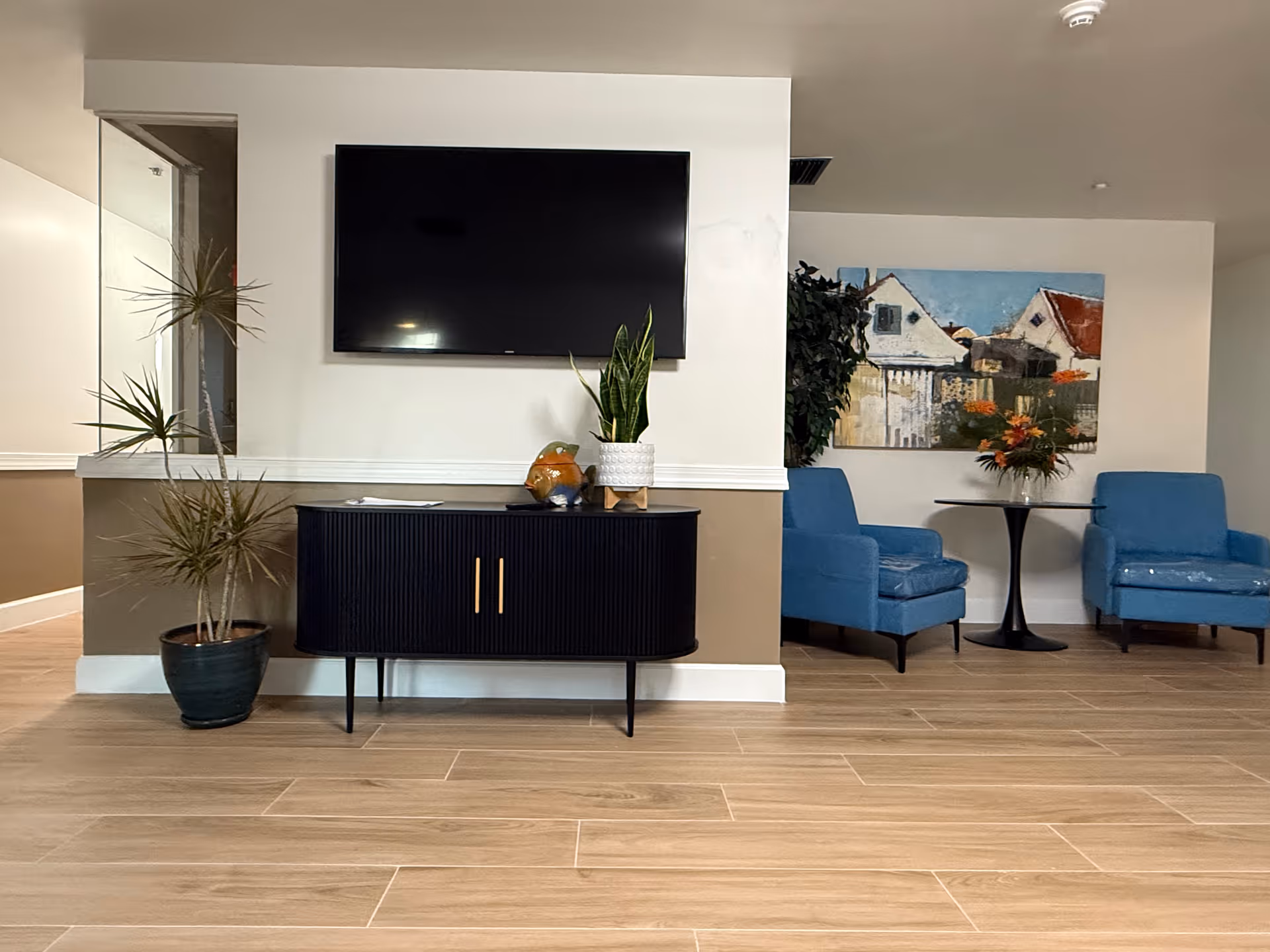 A cozy interior lounge area with a wall-mounted flat screen TV above a black cabinet. There is a potted plant on the floor to the left and another plant on the cabinet. To the right, there are two blue armchairs with a small round black table between them, holding a vase with flowers. A painting of houses hangs on the wall behind the chairs. The floor is covered with light wood-patterned tiles.