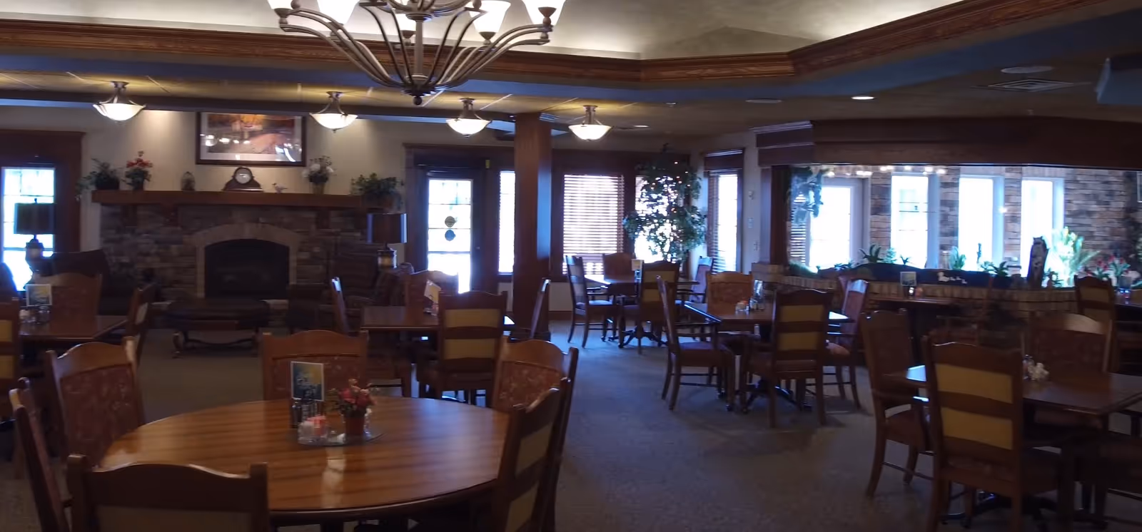Interior view of a dining room with multiple wooden tables and chairs arranged neatly. The room features a stone fireplace with a mantel decorated with plants and a clock. There are large windows with blinds letting in natural light, and a chandelier hanging from the ceiling. The space has a warm and inviting atmosphere with plants and framed artwork on the walls.