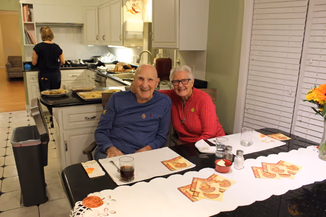 An elderly man and woman sitting closely together and smiling at a dining table set with placemats, napkins, and glasses. Behind them is a kitchen area with a woman preparing food. The room has a warm and welcoming atmosphere.