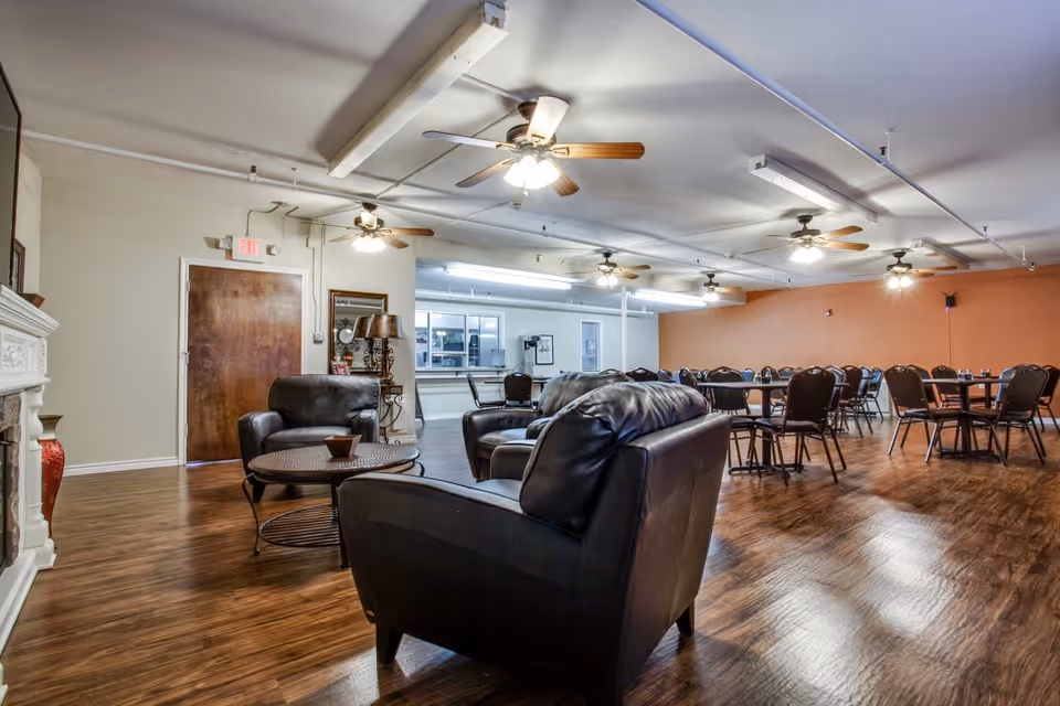 A spacious common area in an assisted living facility featuring several black leather armchairs and a round coffee table in the foreground. In the background, multiple dining tables with chairs are arranged on a wooden floor. The room has ceiling fans with lights and a warm orange accent wall on one side.