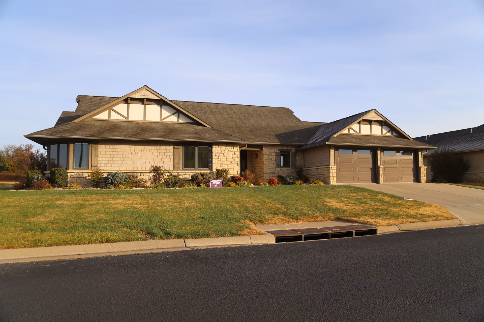 Exterior view of a single-story residential building with a sloped roof, two garage doors, and a well-maintained lawn in front. The building has beige siding with stone accents and decorative wooden trim near the roof peaks. The sky is clear and blue.