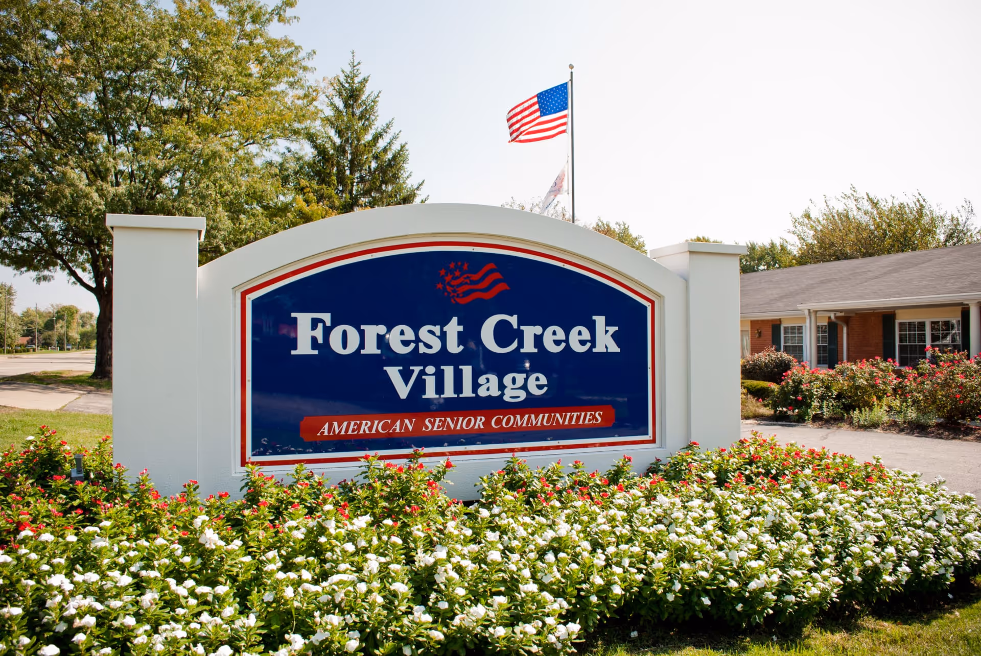 Outdoor view of a sign for Forest Creek Village, an American senior community, surrounded by green bushes with white and red flowers. An American flag is flying on a flagpole behind the sign, with trees and a building visible in the background.
