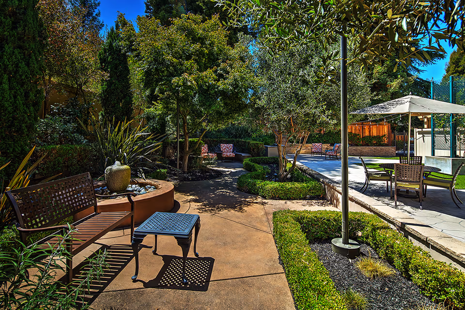 A sunny outdoor garden area with a paved walkway, surrounded by green trees and shrubs. There are several seating options including a wooden bench with a small table, cushioned chairs, and a table with an umbrella. The area is well-maintained with neatly trimmed hedges and landscaping.