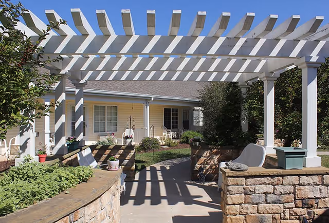 Outdoor patio area with a white pergola casting shadows on the walkway. There are stone walls with plants and flowers, and a yellow building with windows and a porch in the background.