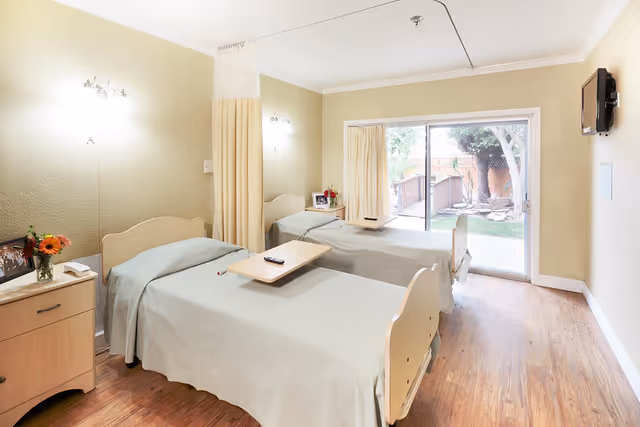 A bright and clean room with two single beds, each with light-colored bedding and a wooden headboard. Each bed has an overbed table with a remote control on it. There is a wooden nightstand with a vase of flowers and a framed photo between the beds. The room has light-colored walls, wooden flooring, wall-mounted lights above each bed, and a large sliding glass door that opens to an outdoor garden area with trees and a small shed. A flat-screen TV is mounted on the wall opposite the beds.