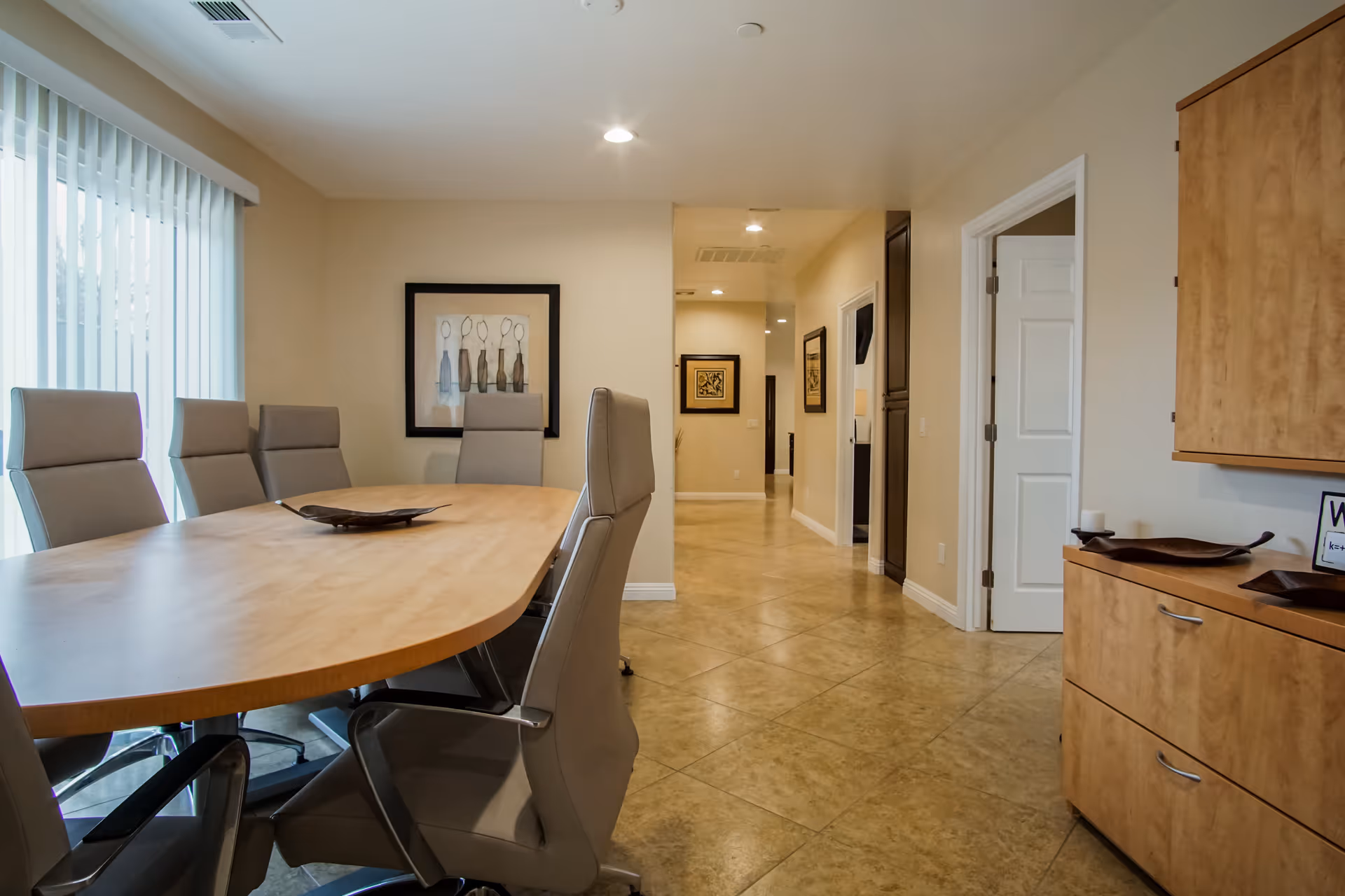 A modern dining room with a long wooden table surrounded by six gray high-back chairs on wheels. The room has beige walls, tiled floor, and a large window with vertical blinds on the left side. There are framed artworks on the walls and a wooden cabinet with decorative items on the right.