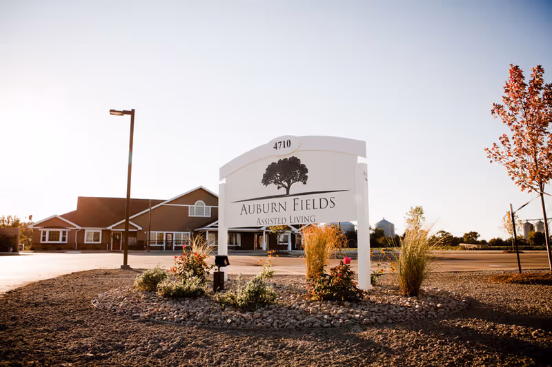 Outdoor view of Auburn Fields Assisted Living facility sign with the building and parking lot in the background under a clear sky during daylight.